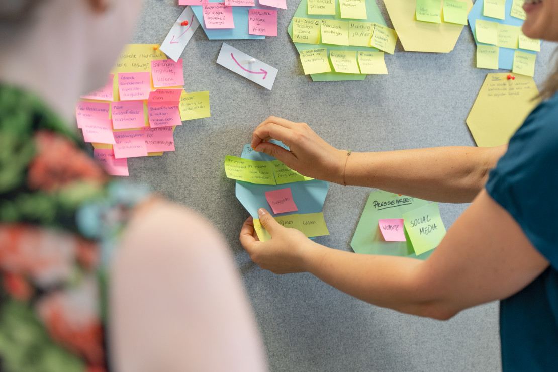 Auf dem Bild sieht man zwei Hände, die etwas an eine Pinnwand pinnen. An der Pinnwand sind viele bunte Postits.